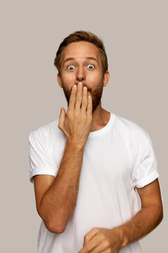 Vertical image of young surprised male in white clothes, covering mouth with hand to be quiet and keep secret, looking at camera with big shocked round eyes. Human emotions and feelings