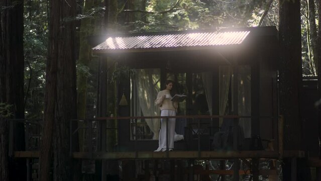 Young woman reading a book and drinking hot tea on a porch of a cozy cabin in the woods. Log cabin in the middle of woods sunny wooden bright mountain cabin rustic house architecture. 