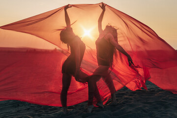 Elegant girls perform dance moves on sand holding up long red fabric against sunset.