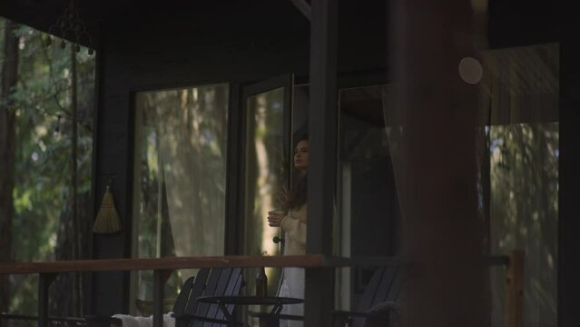 Young woman reading a book and drinking hot tea on a porch of a cozy cabin in the woods. Log cabin in the middle of woods sunny wooden bright mountain cabin rustic house architecture. 