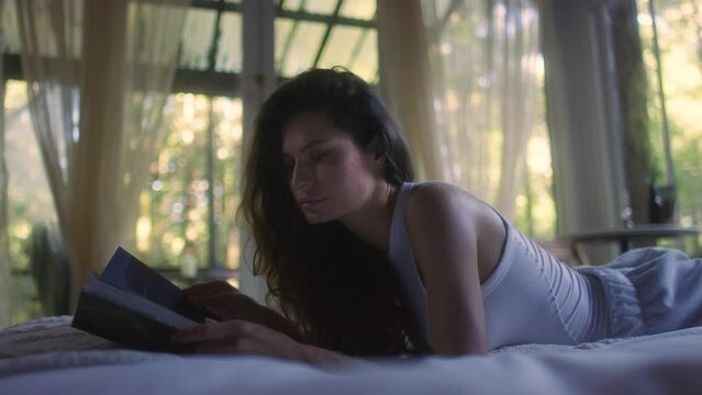 A young woman reads a book on the bed in the bedroom interior. Side view of female enjoying romantic novel lying in bed in the evening. A student read a textbook in the bedroom. Relax time with a book