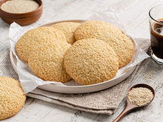Sesame cookies on white ceramic plate with sesame seeds in wooden spoon and coffee glass. Delicious homemade bakery, pastry, dessert or snack. Close up view. White wooden table background, napkin.