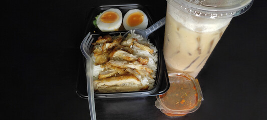 Fried pork with rice and soft boiled egg in a black box Comes with spicy dipping sauce and iced coffee on a black wooden floor. from top view