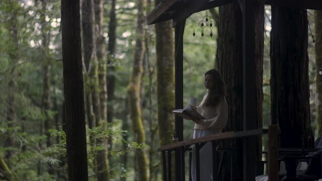 Young woman reading a book and drinking hot tea on a porch of a cozy cabin in the woods. Log cabin in the middle of woods sunny wooden bright mountain cabin rustic house architecture. 