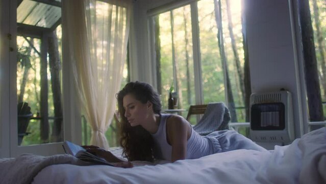 A young woman reads a book on the bed in the bedroom interior. Side view of female enjoying romantic novel lying in bed in the evening. A student read a textbook in the bedroom. Relax time with a book