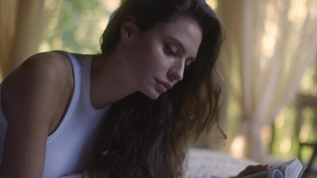 A young woman reads a book on the bed in the bedroom interior. Side view of female enjoying romantic novel lying in bed in the evening. A student read a textbook in the bedroom. Relax time with a book
