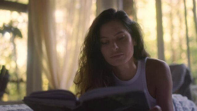 A young woman reads a book on the bed in the bedroom interior. Side view of female enjoying romantic novel lying in bed in the evening. A student read a textbook in the bedroom. Relax time with a book
