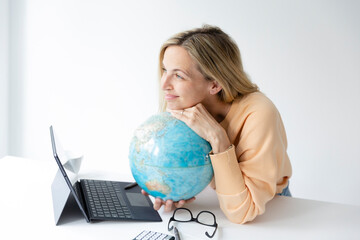 pretty young woman standing at height adjustable table plans her vacation with globe in hand and notebook and is daydreaming