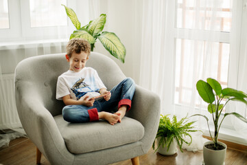 A cheerful boy with a phone in his hands is sitting in a chair and playing a game or watching cartoons.