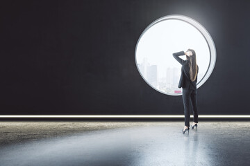 Back view of thoughtful young businesswoman standing in creative modern concrete interior with round window and city view. Mock up place on wall and daylight.
