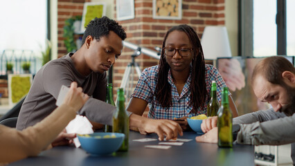 Portrait of cheerful female person playing board games with friends, having fun with cards strategy and competition. Young people enjoying gameplay on table, having beer and snacks.