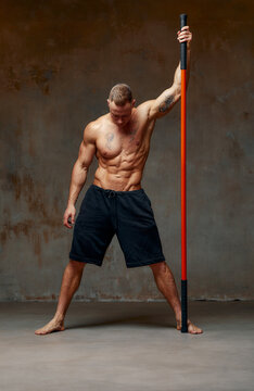 A fighter shirtless in black shorts, stands in a fighting artificially pose and holds a fight stick on studio background.