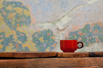 Small red ceramic mug standing on top of wooden table. In the background a worn, colorful wall. Interior, retro.