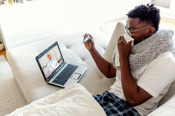 Young man getting online medical help and advice during video call with female doctor and checking symptoms. Ill man lying down at home under blanket and blowing nose, showing thermometer to doctor.