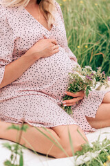 Obraz premium Close-up of a pregnant woman in a pink dress sitting in a meadow on a summer day.Summer,pregnancy and prenatal care concept.