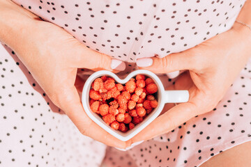 A pregnant woman in a pink dress holds a heart-shaped mug filled with strawberries, hands close-up.Summer,simple living,healthy eating,pregnancy concept.