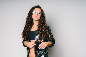 A beautiful young girl with dark curly long hair with glasses in a black shiny shirt looks into the camera. Portrait of a girl waist-high on a white background. Student party. The girl holds her shirt