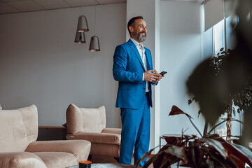 Happy stylish adult man in suit standing in room and holding phone, looking out the window