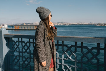 A girl with dark long hair in glasses, in gray coats and a hat looks at the seafront in Istanbul. A student travels during a pandemic. Walks in the autumn in the fresh air. Photo from the back, the - 502538356