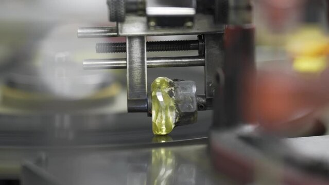 Close up. A large diamond stone rotates as it is cut and shaped using a spinning grinding disk and tang arm by a diamond cutter.
