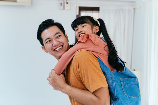 Young father giving his daughter piggyback and smiling in front of house. Happy Asian parenting