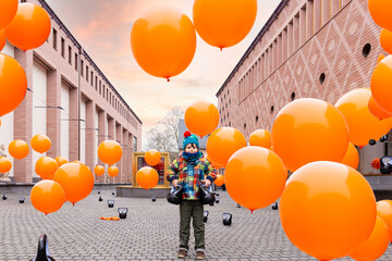 Boy in autumn staying in the street surrounded with orange balloon. Travelling with children in...