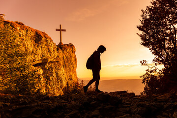 Silhouette of teenager walking on top of the mountain in the sunset, travelling alone, feeling freedom. Cross on the rock. Hiking boy in outdoor adventure.