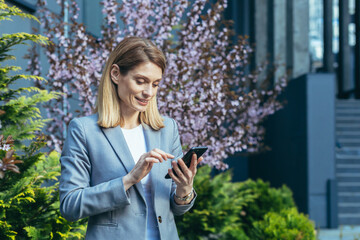Happy business woman uses phone, reads message looking at smartphone screen rejoices and smiles outside office