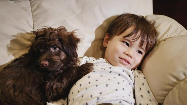 Cute Chinese kid lies on the couch next to a brown puppy, with the TV