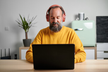Bearded mature man in yellow sweatshirt and wireless headphones sitting at desk with laptop while...