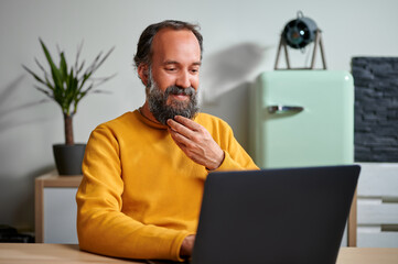 close-up portrait of smiling mature bearded man in yellow sweatshirt sitting at desk with laptop working on remote project at home