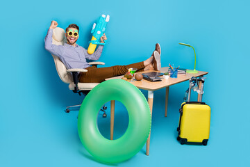 Full length of delighted crazy person sit chair raise fist hold water pistol isolated on blue color background