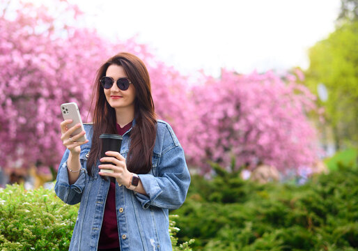 Young beautiful girl standing in sunglasses in the park on a background of pink sakura blossoms in a denim jacket with a mobile phone and coffee. High quality photo