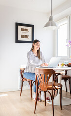 Pregnant woman sitting at table at home using laptop for video call.