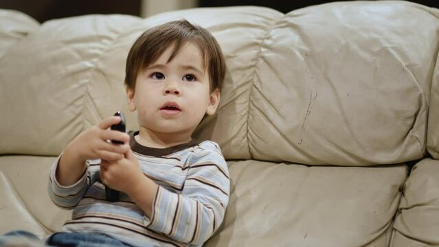 Asian two years old kid watches TV, holds a remote control in his hands