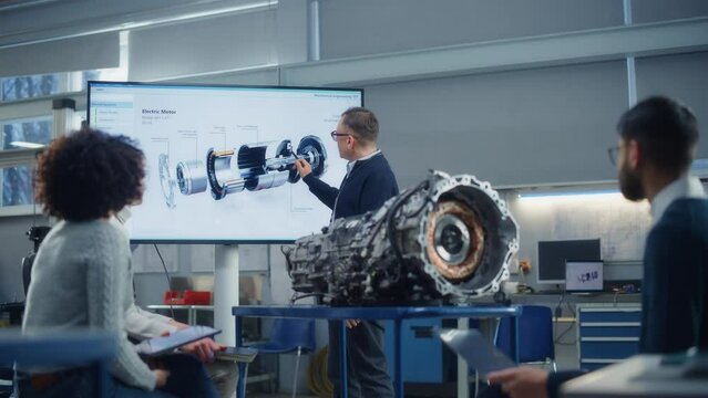 Male Engineer Talks about Work of Turbine Engine to Group of Young Specialists. People Work on Digitalisation of Green Electric Energy. Man Point at Graphics Visualisation on Big Screen and Talk