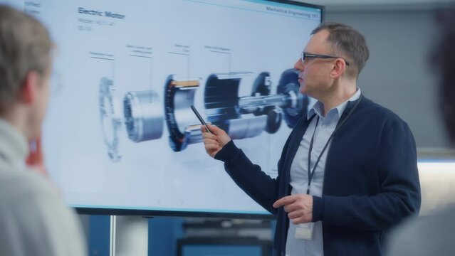 Male Chief Engineer Talks to Young Specialists About Components of the Electric Motor Using Big Screen. Engineering and Green Electric Energy Concept. Portrait view