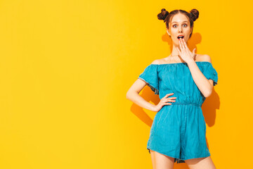 Young beautiful smiling female in trendy summer jeans overalls. Sexy carefree woman with two horns hairstyle posing near yellow wall in studio. Positive model shocked and surprised