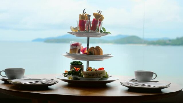Traditional english high tea stand with selection of sweets, cakes, sandwiches, fruit canapes. Afternoon tea tray set with sweet treats on table. Tropical sea and mountains landscape background.