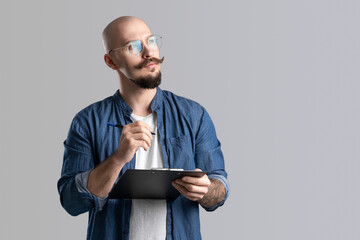 Confident handsome balded beard with musctache man wearing casual clothes holding clipboard isolated over gray background. 