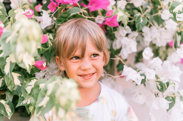 Fototapeta premium portrait of face of a candid little happy smiling five year old blonde kid boy with green eyes in pink and white flower plants in nature. children have fun the summer holidays. bright light and airy