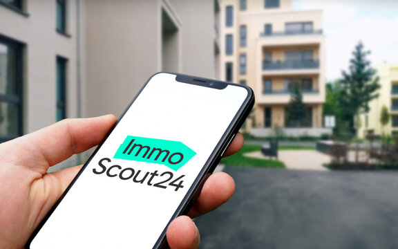 ImmobilienScout24 Logo wird auf einem Smartphone angezeigt, im Hintergrund neu erbaute Mehrfamilienh&auml;user mit Balkonen in einer Stadt, Immo Scout 24, Online-Immobiliensuche, Immobilienb&ouml;rse