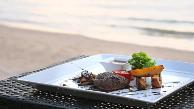 Beefsteak with grilled vegetables dish on white plate on beach table. Filet mignon beef steak, baked potatoes and mushrooms in luxury resort outdoors restaurant. Defocused sandy shore and sea waves.