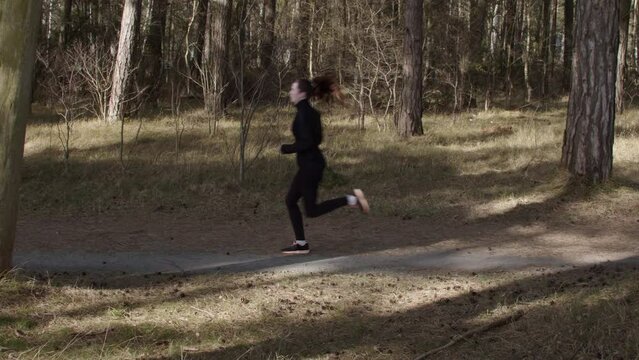 Woman jogging outdoor in forrest on a sunny warm day. Running from right to left in black sportswear. Static wide angle in slow motion. Health motivation. Ystad, Sweden.