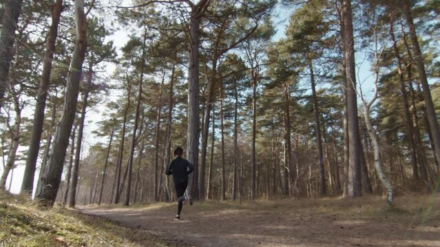 Handheld wide angle shot of young fitness model running past from right to left. Jogging in the forrest among trees and woods in slow motion and 4K. Sunny warm spring day on exercise outdoor trail.