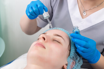 Hands of cosmetologist dripping nourishing oil facial skin of young woman in spa salon.