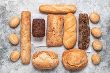 Assortment of various fresh breads on grunge gray background. Varied bakery products. Top view, flat lay. Crispy buns, long loaf, ciabatta and rye bread.