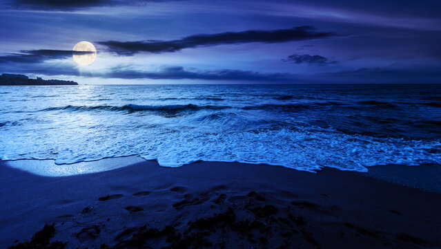 sandy beach at night. beautiful seascape background. calm waves washing the shore. clouds glowing in full moon light. summer vacation and relaxing recreation concept