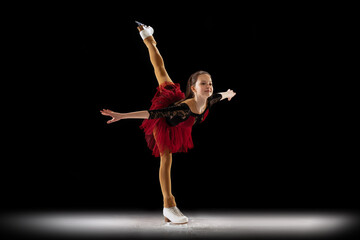 Studio shot of little female figure skater in beautiful stage attire skating isolated on black background in spotlight. Concept of movement, sport, beauty. © master1305
