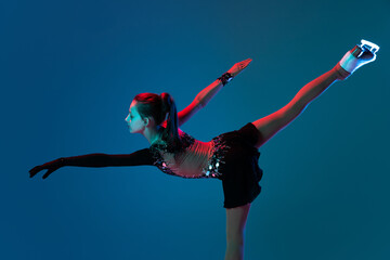 Obraz na płótnie Canvas Dynamic portrait of young girl, female figure skater in black stage dress skating isolated on blue background in neon light. Concept of sport, beauty, active lifestyle.